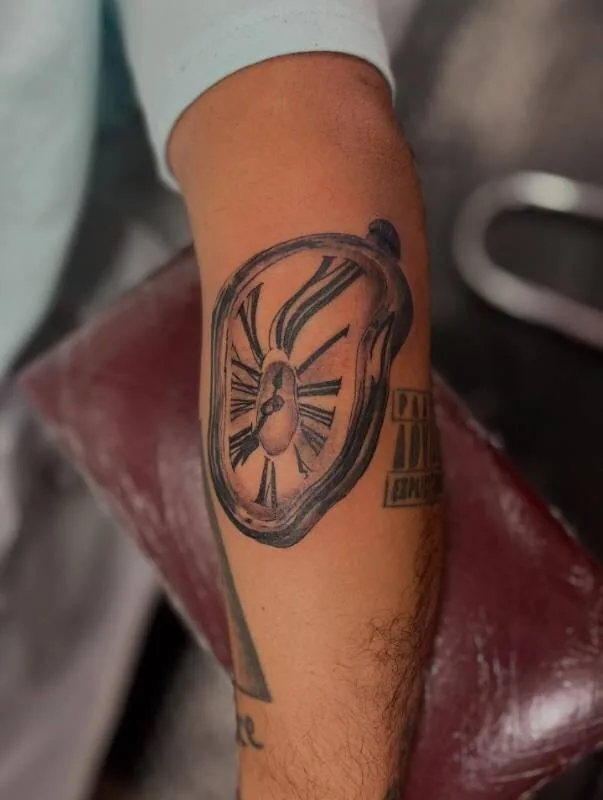 Black and grey surrealist tattoo of a melting clock dripping off the forearm, in tribute to Salvador Dalí, by Franky Sharpz at Wailana Tattoo, Waikiki, Hawaii.